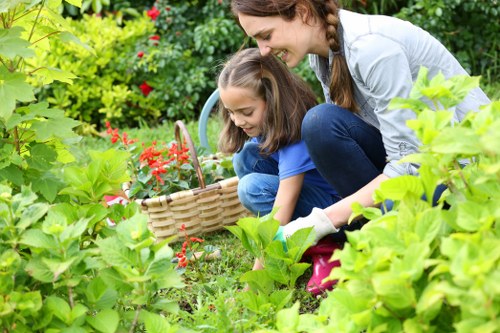A woman and a young girl are engaged in gardening within a lush outdoor space, surrounded by vibrant green foliage, including various shrubs and flowering plants. The woman, dressed in casual clothing, is crouched beside the girl, who is wearing a blue top and jeans, both focusing on planting or tending to the garden bed. They are using a small wicker basket placed on the grass to hold garden tools or harvested flowers. The garden features a well-maintained lawn with dense, healthy grass, and a mix of flowering plants in shades of red and pink, creating a colourful and inviting landscape. In the background, there are taller plants and trees providing a natural border, with daylight illuminating the scene under a clear sky. The overall environment appears peaceful and well cared for, reflecting professional gardening practices by Hedge Trimming Lee, emphasising outdoor maintenance and garden boundary care in a suburban setting near Lee or the South London area.