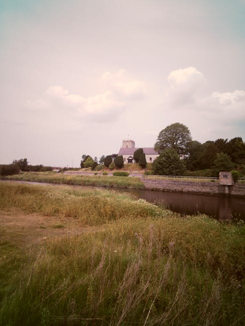 A rural scene featuring a church with a peaked roof and bell tower in the background, surrounded by tall, leafy deciduous trees and well-maintained hedges. In the foreground, there is a grassy field w