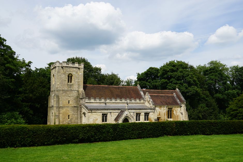 A rural scene featuring a church with a peaked roof and bell tower in the background, surrounded by tall, leafy deciduous trees and well-maintained hedges. In the foreground, there is a grassy field w